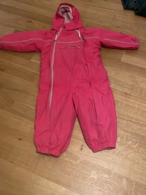 Rosa täckbyxa med huva från Name It - Rosa täckbyxa från Name It med huva och dragkedjor framtill. Overallen har elastiska muddar vid bensluten och en bröstficka med dragkedja. Perfekt för kalla dagar och lek utomhus. Fodrad insida för extra värme.