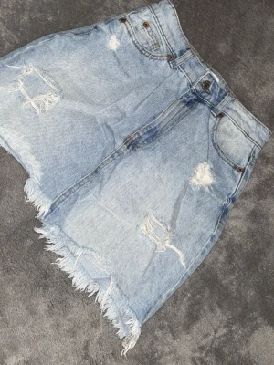 Ljusblå jeanskjol med fransad kant - Säljer en ljusblå jeanskjol från Levi's med slitna detaljer och fransad kant. Kjolen har klassiska fem fickor och råa kanter nertill för en avslappnad look. Perfekt för dig som gillar en trendig och edgy stil.