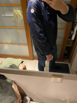 Mörkblå vindjacka från Moncler - Snygg mörkblå vindjacka från Moncler med dragkedja framtill och två sidofickor med tryckknappar. Jackan har Moncler-logga på ärmen och en klassisk, enkel design som passar till jeans. Tillverkad i lätt och vindtätt material är som ny har använt den några gånger.