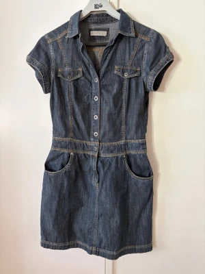 Jeansklänning  - Snygg jeansklänning från Lindex med korta ärmar, krage,  knappar och dragkedja i gylfen framtill. Klänningen har två bröstfickor med lock, stora sidofickor och två bakfickor. Klassisk blå denim med kontrastsömmar och en figurnära passform. Liten 38 mer lik en 36’a