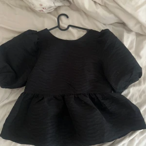 Svart topp med puffärmar och knyt - Trendig svart topp med voluminösa puffärmar och knyt i ryggen. Toppen har en lös passform och en lätt rynkad nederdel som ger en cool siluett. Perfekt för dig som vill sticka ut med en statement-topp. Ny pris 600