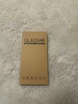 GLASS PRO skärmskydd härdat glas - Säljer ett GLASS PRO skärmskydd i härdat glas. Förpackningen är beige och skyddet är genomskinligt. Perfekt för att skydda din mobilskärm mot repor och stötar. Enkel installation med tydliga instruktioner på baksidan av förpackningen. iPhone 14 Pro 