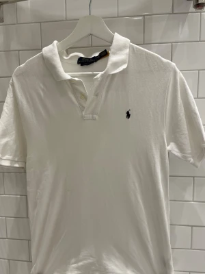 Vit pikétröja från Ralph Lauren - Klassisk vit pikétröja från Ralph Lauren med korta ärmar och den ikoniska broderade loggan på bröstet. Tröjan har krage och knappar framtill. Perfekt för en clean och stilren look.