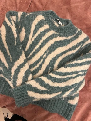 Stickad tröja med zebramönster - Säljer en fluffig stickad tröja med zebrainspirerat mönster i grönt och vitt. Tröjan har rund halsringning, långa ärmar och ribbade muddar. Perfekt för dig som vill ha en trendig och mysig look.