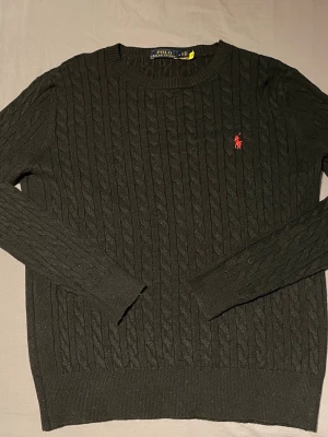 Svart kabelstickad tröja Polo Ralph Lauren -  Kabelstickad svart tröja från Polo Ralph Lauren med rund halsringning och lång ärm. Tröjan har det klassiska röda broderade logotypen på bröstet och ribbade muddar vid ärmslut och nederkant. Perfekt för en sopistikerad och avslappnad look. Storlek M fast passar bäst på S. Säljer pågrund av att den satt för liten på mig julklapp från början. Kom privat för bilder. Pris kan diskuteras 