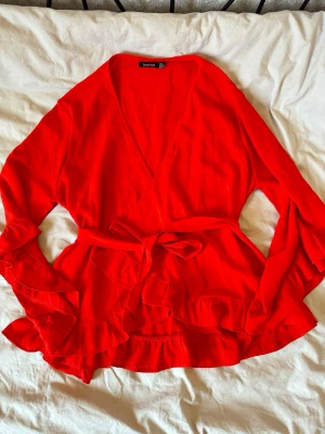 Röd omlottblus med volanger Boohoo - Snygg röd omlottblus från Boohoo med långa ärmar och volangdetaljer vid ärmslut och nederkant. Blusen har ett knytband i midjan och djup V-ringning. Perfekt för dig som vill sticka ut med färg och trendiga detaljer.
