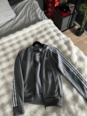 Grå Adidas ziptröja med vita ränder - Säljer en grå ziptröja från Adidas med klassiska vita ränder längs ärmarna och Adidas-logga på bröstet. Tröjan har dragkedja framtill, ribbade muddar och är tillverkad i ett mjukt, sportigt material. Perfekt för en avslappnad streetwear-look. Använd 2 gånger , finns att köpa byxor till samma färg , nypris för båda är 1400 kr 