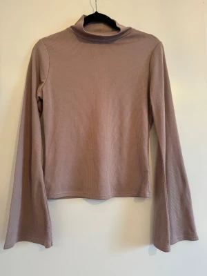Beige ribbad polotopp med lång ärm - Snygg beige ribbad polotopp med lång ärm och hög krage. Toppen har en smickrande passform och är tillverkad i ett mjukt, stretchigt material som känns skönt mot huden. Perfekt att styla med jeans eller kjol för en clean look.