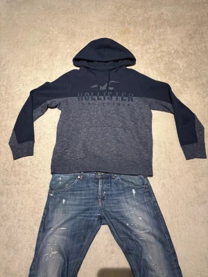 Mörkblå Hollister hoodie med logga - Snygg mörkblå hoodie från Hollister med stor logga och texten 'Hollister California' på bröstet. Hoodien har dragsko i huvan och är tillverkad i mjukt bomullsmaterial. Perfekt för en avslappnad och trendig stil.