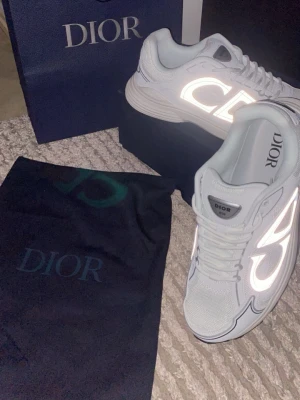 Vita Dior sneakers med reflekterande detaljer - Säljer ett par vita Dior sneakers med stora reflekterande loggor på sidan. Skorna har en chunky sula och är tillverkade i mesh och läder för en modern och exklusiv look. Perfekta för dig som vill sticka ut med stilrena och lyxiga sneakers.