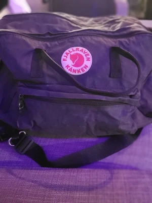 Svart Kånken ryggsäck Fjällräven - Klassisk svart Kånken ryggsäck från Fjällräven med det ikoniska röda logotypmärket framtill. Ryggsäcken har ett stort huvudfack, en dragkedjeförsedd framficka och justerbara axelremmar. Tillverkad i slitstarkt syntetmaterial som tål vardagens äventyr.