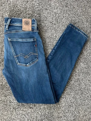 Blå slim fit jeans från replay - Säljer ett par blå slim fit jeans från replay med snygga slitningar och klassisk femficksmodell. Jeansen har en skinnpatch bak i midjan och kontrastsömmar på bakfickorna. Tillverkade i mjuk denim med stretch för extra komfort.