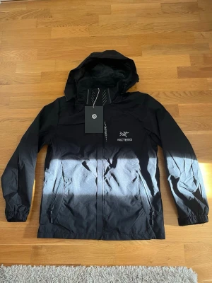 Svart vindjacka från Arc'teryx - Snygg svart vindjacka från Arc'teryx med huva och dragkedja framtill. Jackan har två sidofickor med dragkedja, justerbar huva och elastiska muddar vid ärmsluten. Tillverkad i lätt och slitstarkt material, perfekt för blåsiga dagar.