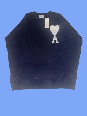 Mörkblå stickad tröja från Ami - Snygg mörkblå stickad tröja från Ami med rund halsringning och ribbade muddar. På bröstet finns ett vitt hjärta och bokstaven A som ger en unik touch. Perfekt för dig som gillar stilrena detaljer och vill ha något trendigt i garderoben.