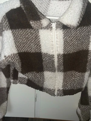 Brunvitrutig teddyjacka med dragkedja - Mysig teddyjacka i brun och vit rutigt mönster. Jackan har en croppad passform, stor krage och dragkedja framtill. Tillverkad i fluffigt teddy-material som ger en varm och trendig look. Perfekt för dig som gillar streetstyle och vill ha något riktigt skönt.🤍🤎den är i storkek S