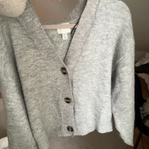 Ljusgrå kofta med knappar - Mysig ljusgrå kofta med v-ringning och stora bruna knappar framtill. Koftan har en avslappnad passform och är gjord i ett mjukt, stickat material. Perfekt att slänga på sig över en t-shirt eller linne. Jättefint att ha som off-shoulder, hör gärna av om ni har frågor!
