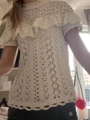 Beige spetsblus med volang från Zara - Supersöt beige spetsblus från Zara med korta ärmar och broderad volangdetalj över bröstet och axlarna. Blusen har hög krage med volangkant och ett vackert hålmönster i hela tyget. Perfekt för dig som gillar romantisk och vintageinspirerad stil.