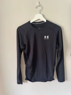 Svart långärmad träningströja Under Armour - Svart långärmad träningströja från Under Armour med vit logga på bröstet och nacken. Tillverkad i ett stretchigt funktionsmaterial som andas, perfekt för intensiva träningspass eller löpning. Tight passform och rund halsringning.