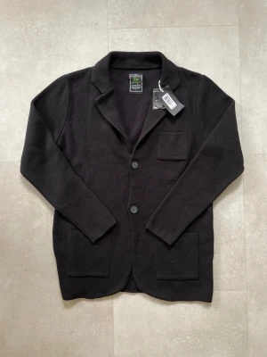 Svart stickad kavajkofta  - Snygg svart stickad kavajkofta med två knappar framtill, tre fickor och klassisk krage. Perfekt för dig som vill ha en avslappnad men ändå stilren look. Materialet är mjukt och följsamt, vilket gör den bekväm att bära hela dagen.