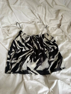 Zebramönstrad linnetopp med tunna band - Säljer en cool zebramönstrad linnetopp med tunna axelband och knytning framtill. Toppen är svart och vit, har en luftig passform som passar perfekt till varma dagar. Materialet känns lätt och bekvämt mot huden. Perfekt som en lite finare tröja eller en tröja som går att klä ner. Justerbara band. 