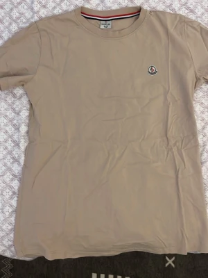 Beige Moncler t-shirt i bomull - Snygg beige t-shirt från Moncler med klassisk rund hals och korta ärmar. Liten Moncler-logga på bröstet och diskret design. Tillverkad i mjuk bomull som känns skön mot huden. Perfekt till jeans eller shorts för en clean look.