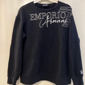 Svart sweatshirt från Emporio Armani - Säljer en svart sweatshirt från Emporio Armani med vit logotyp på bröstet. Tröjan har långa ärmar och en rund halsringning. Perfekt för en stilren och bekväm look.