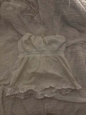 Vit broderad topp med volang - Söt vit topp med tunna axelband och broderade blommor. Modellen är ärmlös och har volangkant nertill samt smock i midjan för en feminin siluett. Perfekt för varma dagar. Vid inresse kan jag skicka fler bilder!🙌