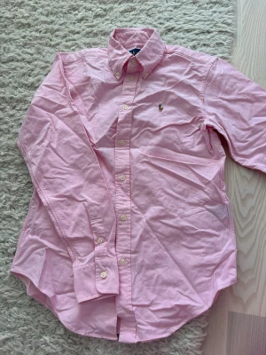 Rosa skjorta från Ralph Lauren - Klassisk rosa skjorta från Ralph Lauren med button-down krage och broderad logga på bröstet. Skjortan har långa ärmar, vita knappar och är tillverkad i ett mjukt bomullstyg. Perfekt för en stilren och avslappnad look.