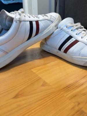 Vita Moncler sneakers med ränder - Snygga vita sneakers från Moncler med klassiska röda och mörkblå ränder på sidan. Skorna har låg profil, rund tå och är tillverkade i läder med Moncler-logga på sidan och hälen. Perfekt för dig som gillar stilrena och exklusiva sneakers.