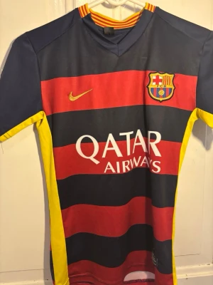 Barcelona Neymar JR matchtröja (2015) - Säljer en FC Barcelona matchtröja från Nike med Neymar JR och nummer 11 på ryggen. Tröjan har breda röda och mörkblå ränder, gula detaljer och klubbens emblem på bröstet. Materialet är lätt och andas, perfekt för fotboll eller träning.