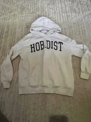 Vit hoodie med tryck från HOB.DIST - Vit hoodie med svart texttryck över bröstet från lager 157. Klassisk huva och ribbade muddar vid ärmslut och nederkant. Perfekt för en chill och avslappnad stil.