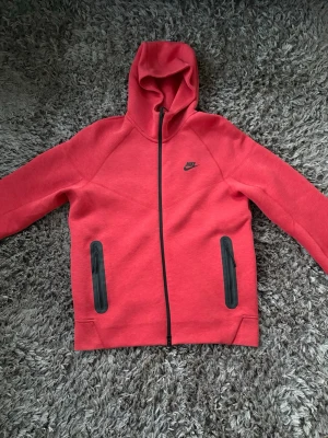 Röd Niketech med dragkedja - Snygg röd hoodie från Nike med hel dragkedja och två svarta dragkedjefickor framtill. Klassisk huva och diskret Nike-logga på bröstet. Perfekt för en sportig och avslappnad stil.