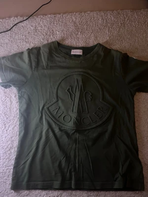 Olivgrön Moncler t-shirt med logga - Snygg olivgrön t-shirt från Moncler med stor, upphöjd logga på bröstet. Klassisk rund halsringning och korta ärmar. Tillverkad i mjuk bomull som känns skön mot huden. Perfekt för dig som gillar streetwear och exklusiva märken.
