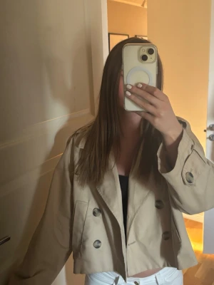 Kort beige trenchcoat med dubbelknäppning - Snygg kort trenchcoat i beige med klassisk dubbelknäppning och breda slag. Jackan har axelklaffar och är tillverkad i ett lätt material som passar perfekt till vår och höst. Stilren och trendig modell som är enkel att matcha.