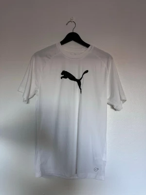 Vit Puma t-shirt med svart logga - Snygg vit t-shirt från Puma med den klassiska svarta loggan på bröstet. T-shirten har rund halsringning och korta ärmar. Tillverkad i ett lätt och bekvämt material som passar perfekt till en aktiv livsstil.