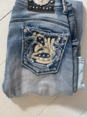Ljusblåa  jeans med broderi - Säljer ett par ljusblåa jeans från tror  Miss Me men inte säker med coolt broderi och slitningar på bakfickorna. Jeansen har dekorativa nitar och en unik design som verkligen sticker ut. Perfekta för dig som gillar detaljer och en trendig look. Bootcut o lågmidjade
