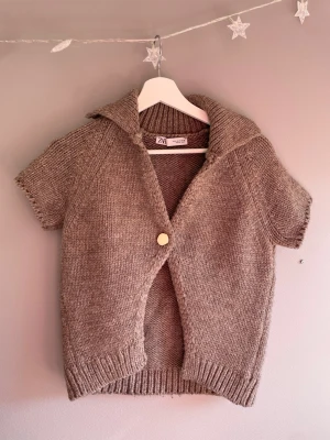 Brun stickad kortärmad kofta från Zara - Stickad brun kofta från Zara med korta ärmar och bred krage. Koftan har ett stort beige knapp framtill och ribbstickade detaljer vid ärmslut och nederkant. Perfekt att slänga över en t-shirt för en chill look.