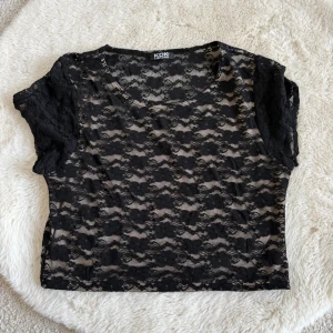 Svart spetsblus från KDK London (XS) - Säljer en svart, kortärmad spetsblus från KDK London. Blusen är genomskinlig med blommigt spetsmönster och har en rund halsringning. Perfekt för att skapa en cool och trendig look.