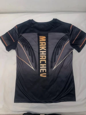 Islam Makhachev tshrit - Cool svart t-shirt med Makhachev-tryck på ryggen och Crypto.com-logga på bröstet. T-shirten har korta ärmar och ett futuristiskt mönster i grått och orange. Materialet är polyester och passar perfekt för dig som gillar sportiga och moderna plagg.