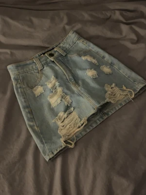 Blå jeanskjol med slitningar - Cool kort jeanskjol i ljusblå denim med flera slitningar och råa kanter. Kjolen har klassiska fickor fram och bak, bälteshällor och knappstängning. Perfekt för dig som gillar en edgy och trendig look.