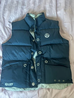 Vändbar dunväst  - Snygg reversible dunväst från märket North Sails med ena sidan i mörkblått och andra i ljusgrått. Västen har hög krage, tryckknappar, dragkedjor och praktiska fickor på båda sidor. Märkeslogga på bröstet och coola detaljer vid fickorna. Perfekt för lager-på-lager och streetwear. Unisex