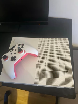 Xbox One S vit spelkonsol med handkontroll - Säljer en Xbox One S i snygg vit design med tillhörande vit handkontroll som har rosa detaljer. Konsolen har en stilren, rektangulär form och är tillverkad i plast med ventilerad ovansida för bra kylning. Perfekt för gaming och streaming.