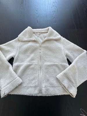 Vit teddyjacka med dragkedja - Mysig vit teddyjacka i storlek S med dragkedja framtill och två stora fickor. Jackan har en klassisk krage och är gjord i fluffigt, mjukt material som håller dig varm. Perfekt för kyliga dagar och enkel att matcha med olika outfits.