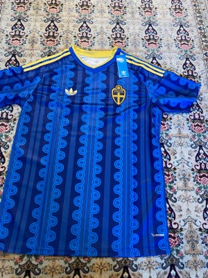 Blå Sverige bortatröja - Säljer en blå fotbollströja med Sveriges landslagsemblem på bröstet. Tröjan har ett unikt mönster i ljusblått, gula detaljer vid krage och axlar samt Adidas logga. Materialet är lätt och andas, perfekt för träning eller match.