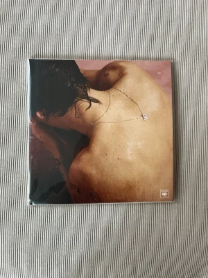 Harry Styles vinylalbum - Vinylalbum med omslag i rosa och beige toner, visar en person med blött hår och halsband. Baksidan har låtlista och ett mindre foto. Albumet är fyrkantigt och tillverkat av klassiskt vinylmaterial.