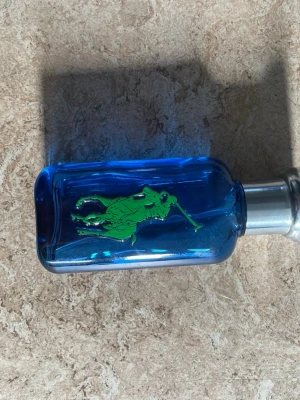 Blå parfymflaska från Ralph Lauren - Snygg parfymflaska i blått glas från Ralph Lauren med en grön polospelare på framsidan. Flaskan har en silverfärgad spraykork och en rektangulär form. Perfekt för dig som gillar sportiga och fräscha dofter.