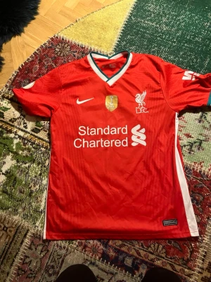 Liverpool matchtröja Nike röd - Snygg röd Liverpool FC matchtröja från Nike med vita detaljer och klubbmärke på bröstet. Tröjan har V-ringning med vit och grön kant, tryckt sponsorlogga och korta ärmar. Perfekt för fotbollsfans som vill visa sitt stöd på matchdag.
