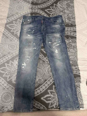 Blå distressed jeans från Dsquared2 - Säljer ett par blå jeans från Dsquared2 med coola slitningar och distressed detaljer. Klassisk femficksmodell med rakt ben och snygg tvätt. Perfekta för dig som gillar en avslappnad och trendig stil.