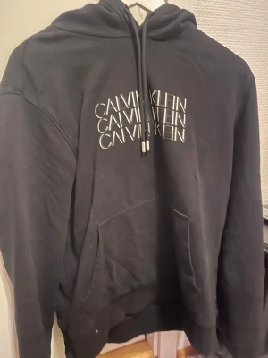 Svart hoodie från Calvin Klein - Svart hoodie från Calvin Klein med vit logga tryckt flera gånger på bröstet. Klassisk känguruficka framtill, dragsko i huvan och mjukt material. Perfekt för en avslappnad och trendig stil.