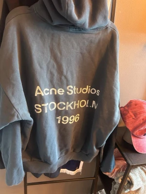 Mörkgrå hoodie Acne Studios - Cool mörkgrå hoodie från Acne Studios med stor huva och tryck på ryggen: 'Acne Studios STOCKHOLM 1996'. Oversized passform och mjukt material som känns riktigt skönt. Perfekt för dig som gillar streetstyle och vill ha något trendigt.